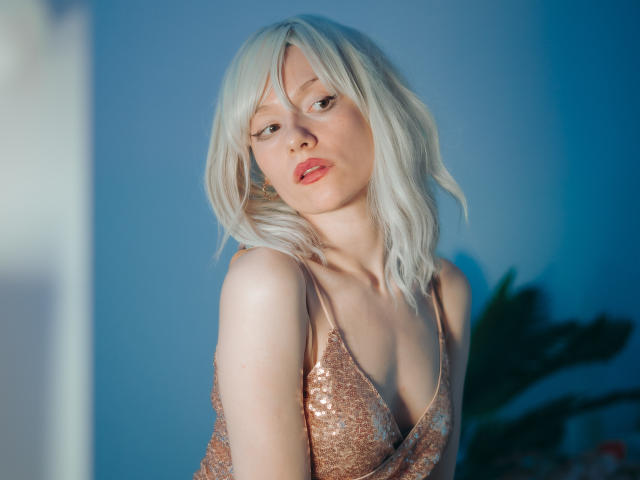 Brenda Lovex mit platinblondem Haar, in einem glitzernden, kupfarbenen Kleid, das ihre kleinen Brüste zeigt, mit heller Haut, roten Lippen und leicht geneigtem Kopf, auf einem Foto mit blauem Licht..