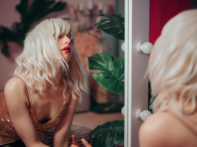 Brenda Lovex mit platinblondem Haar, rotem Lippenstift und einem glitzernden goldfarbenen Kleid, lehnt sich vor und betrachtet sich in einem Spiegel mit runden Glühbirnen, wobei ihre Brüste zu sehen sind..