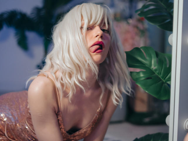BrendaLovex with blonde hair, tongue out, wearing a sparkling pink dress, has her tits slightly visible, standing next to a large green plant.