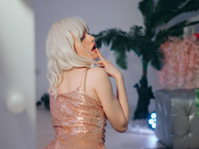 BrendaLovex with blonde wig, wearing a sparkly pink dress, has her back to the camera, biting her finger, showing off her bare shoulders and ass.