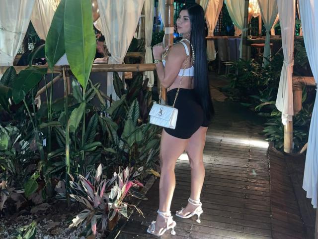 Sarah, con lunghi capelli neri, reggiseno bianco, minigonna nera, tacchi alti bianchi e borsa bianca, in piedi su un sentiero di legno, guarda indietro, mostrando il sedere e la pelle abbronzata..