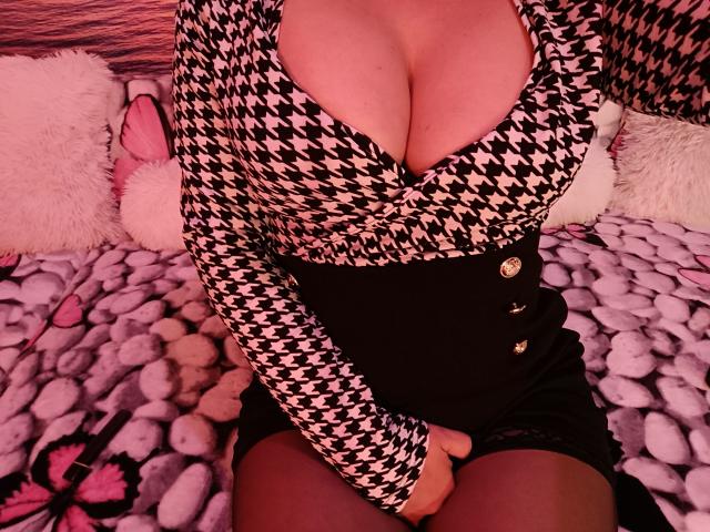 AmberSunshine with big tits in a houndstooth top, black skirt with gold buttons, sitting on a pink and white textured blanket, hands on her thighs.