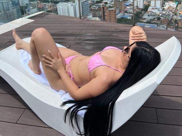 Sarah, com longos cabelos pretos, biquíni rosa e óculos de sol, relaxando em um balanço branco em um deck de madeira, com as pernas levantadas, as mãos sobre as coxas, exibindo suas nádegas e seios..