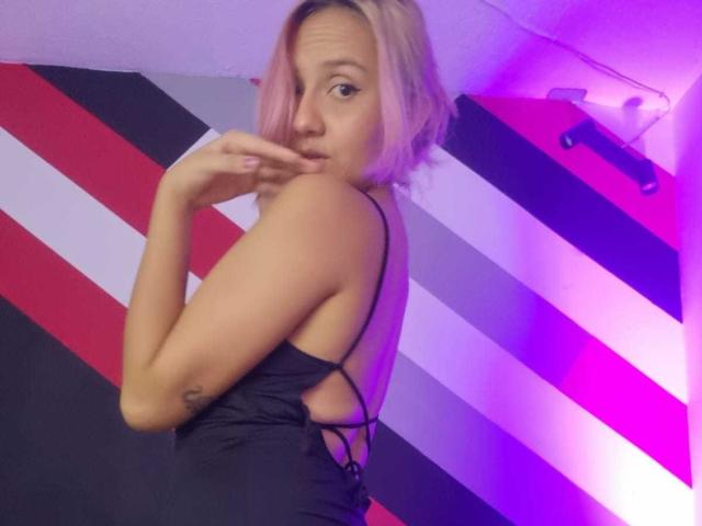 AlterEgoSecond con capelli rosa, vestito nero, che mostra la schiena, con un dito in bocca, tatuaggio sul braccio sinistro, che guarda oltre la spalla, illuminazione viola e rosa..