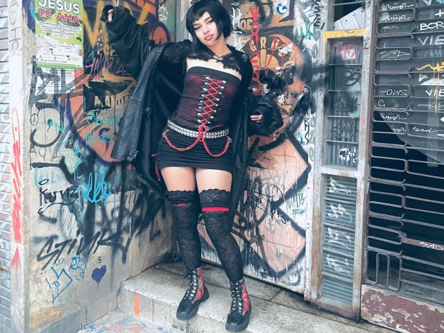 BatiDark with black bob, red-laced corset, black mini dress, lace thigh-highs, and black boots, standing in graffiti-covered alleyway, wearing open black jacket, holding black bag.