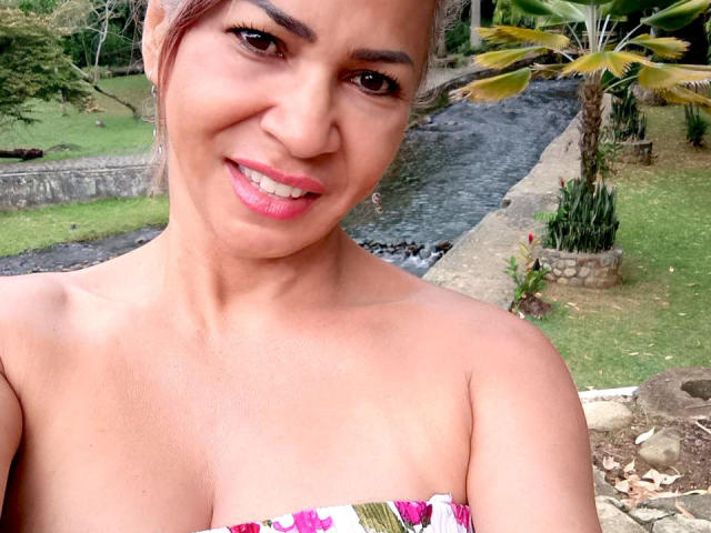 Brigitte Walsh, com pele marrom clara, batom rosa e olhos castanhos, sorrindo para a câmera, mostrando seus ombros nus e um top colorido sem alças, com seios pequenos e visíveis..