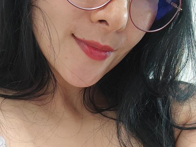 AlmaSil has pale skin, dark wavy hair, and pink glasses. She's smiling with red lipstick, showing her soft, cute tits in a white top.