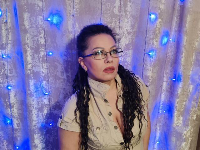 AngellaFly with long curly black hair, glasses, and a low-button shirt, exposing her tits, standing against a blue-lit, metallic curtain.
