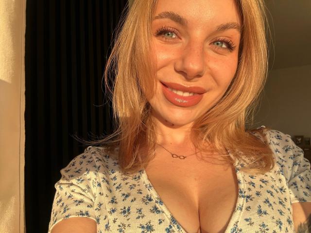 SlaviaH-hot with blonde hair, blue eyes, and a smile, wearing a floral dress, showcasing her big tits and a heart necklace, illuminated by warm sunlight.
