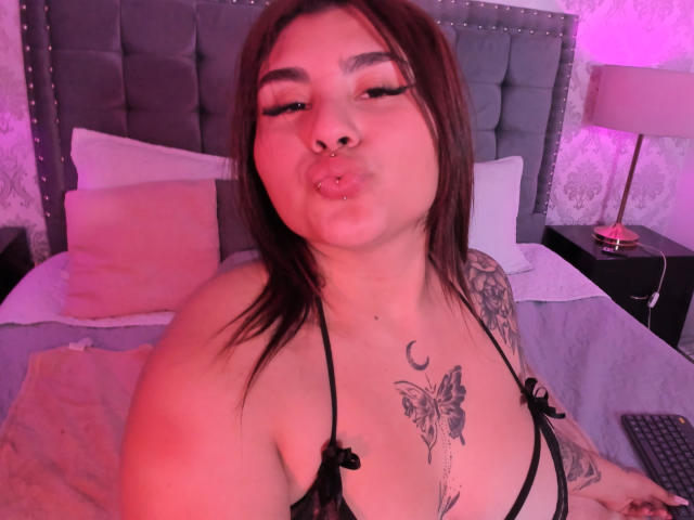 KatiaRodriguez with pouty lips, brown hair, tattoos on chest and arm, wearing black lingerie, pink and purple lighting, gray tufted headboard, and pink pillow in background.