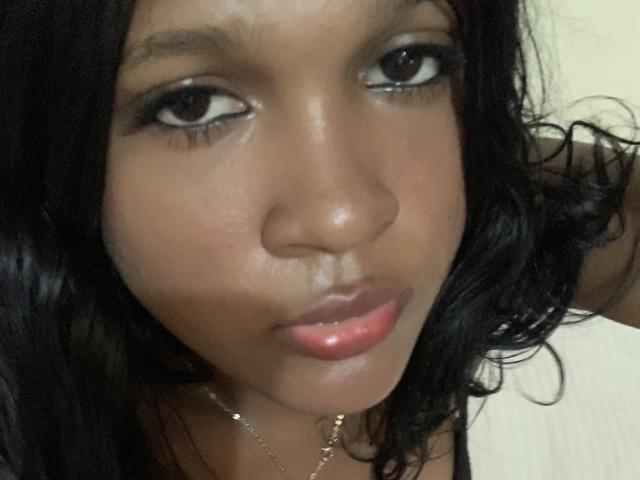 SexyHabibi has dark skin, shiny black wavy hair, and full pink glossy lips. She's looking up with big brown eyes, wearing subtle makeup and a gold necklace.