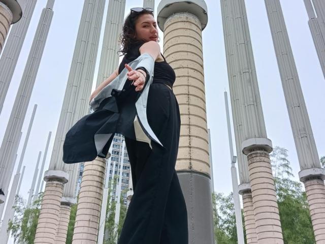 ElizaCanterville has curly brown hair, wearing a black sleeveless top and black pants, holding a blue jacket, standing in front of tall cylindrical pillars, looking back confidently.