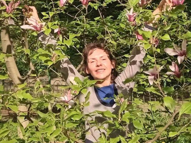 GrostFlow with arms raised, smiling, surrounded by green leaves and pink magnolia flowers. Wearing a gray long-sleeve shirt and blue scarf, standing in a sunny, leafy garden.