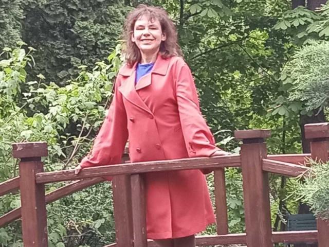 GrostFlow, middle-aged woman with brown curly hair, wearing a red coat, blue shirt, and black pants, standing on a wooden bridge, smiling, hands on railings.