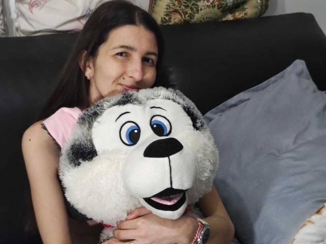 DarrylAndCelia with a fluffy grey and white stuffed dog, hugging it. DarrylAndCelia has long brown hair, wearing a pink shirt, smiling, and looking at the camera.
