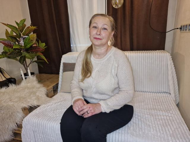 AvroraRosiye, middle-aged woman, sitting on white ribbed couch, wearing fluffy beige sweater, black pants, blonde ponytail, pink lipstick, small tits, hands on knees, brown curtains in background.