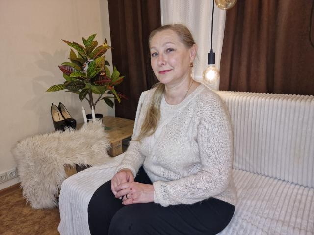 AvroraRosiye has pale skin, blonde hair in a ponytail, wearing a white knit sweater, black pants, and a subtle red lipstick, sitting on a white couch with a furry pillow.