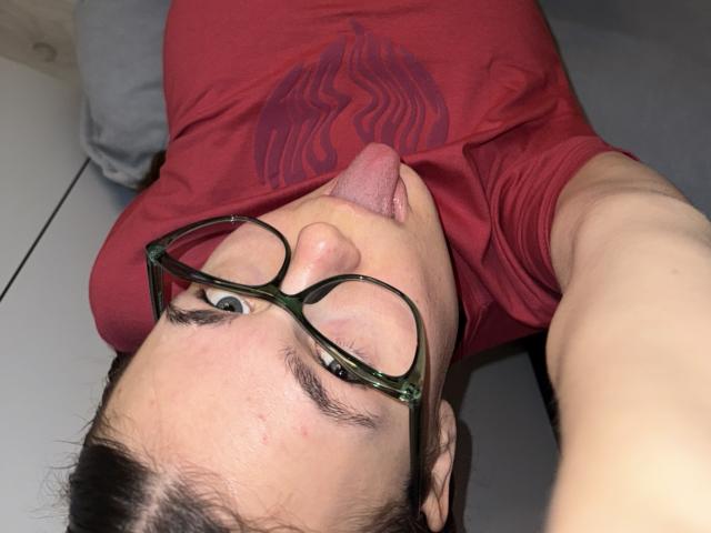LinaStones, wearing green glasses, red shirt, and sticking out tongue, lying on grey surface, taking selfie. Her face is close to camera, with slight pimple on nose.