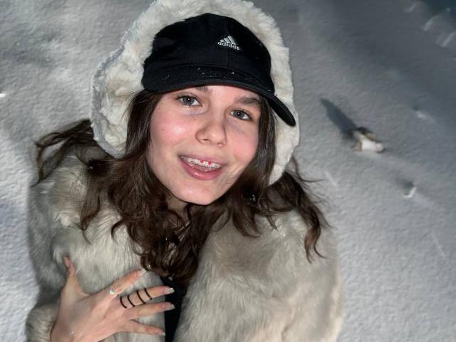 LinaStones with brown hair, wearing a black Adidas cap and a furry coat, smiling in a snowy setting. Her hand shows a ring. She's got a cute, slightly goofy face.