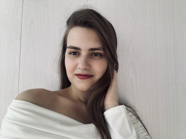 VikkiGorgeous, com cabelos castanhos longos, camisa branca com um ombro descoberto, batom vermelho e um sorriso discreto, com a mão no cabelo, pele clara e expressão suave, olhando ligeiramente para a direita..