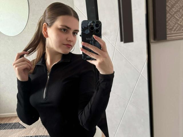 VikkiGorgeous mit zurückgekämmtem, braunem Haar, trägt eine schwarze Jacke mit Reißverschluss und macht ein Selfie mit ihrem schwarzen Handy. Sie zeigt ein leichtes Lächeln und hält ihr Haar mit ihrer linken Hand..