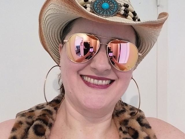 LiliChattePoilue with pinkish skin, smiling, wearing a cowboy hat, reflective pink aviator sunglasses, large gold hoop earrings, leopard print top, and pink lipstick.