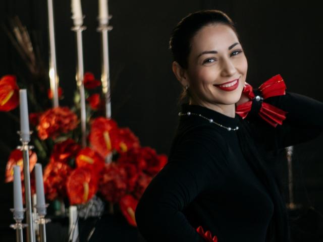 LiluGrants with red lipstick, smiling, wearing a black dress with red bows, has her hand on her head, standing next to red flowers and white candles.