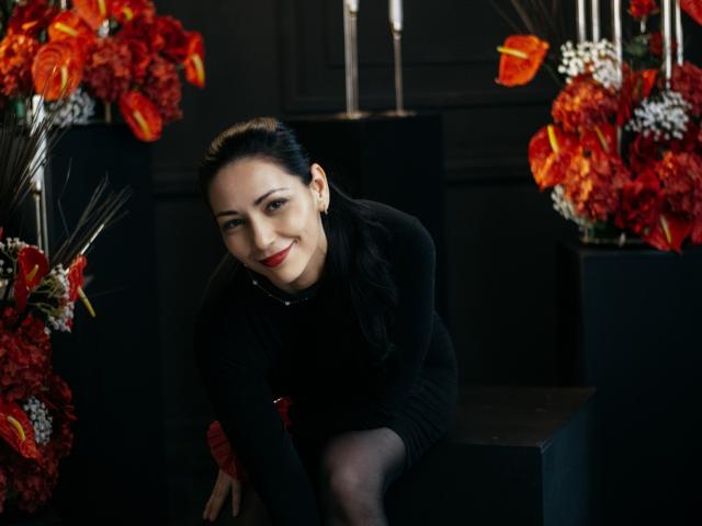 LiluGrants with dark hair, red lipstick, and black sweater, smiling at camera, surrounded by red and orange flowers, sitting on dark surface, with candles in background.