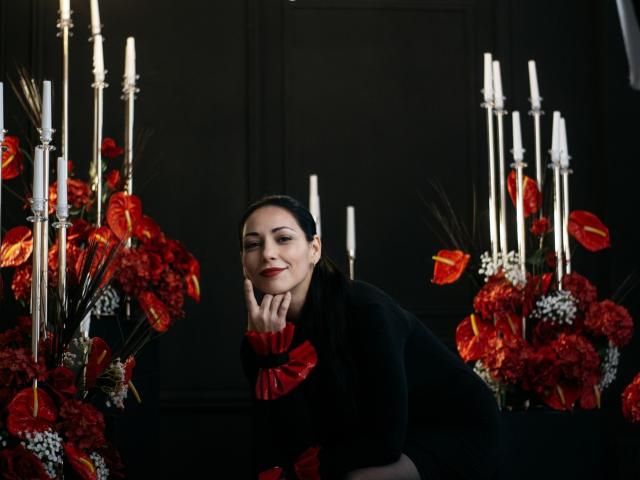 LiluGrants with dark hair, black dress, red flower accent, leaning on hand, smiling, surrounded by tall white candles and red, orange, and white flowers.