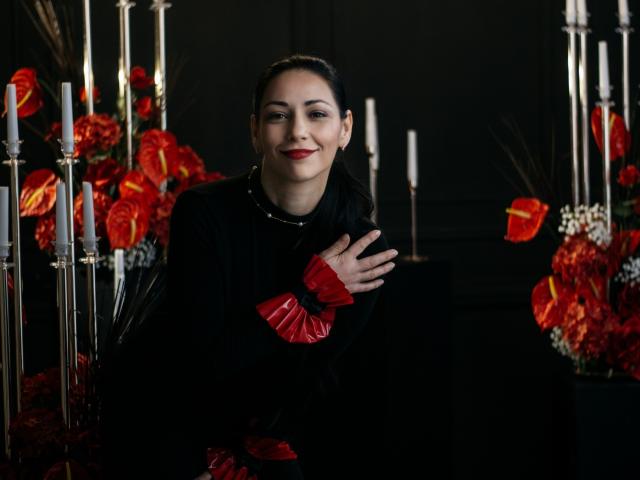 LiluGrants, with red lips and black dress, holding chest, red cuffs, dark hair, smiling, surrounded by red flowers and white candles, looking sexy and confident.