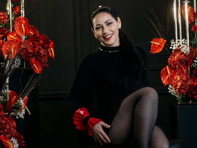 LiluGrants with dark hair in a ponytail, black dress, red wrist cuffs, and black pantyhose, smiling with red lips, sitting with one leg up, surrounded by red and orange flowers.