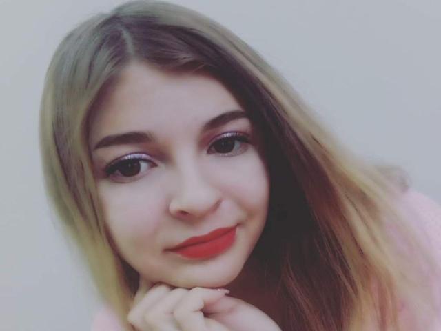 RoxannaSonia has long blonde hair, red lipstick, and a cute smile, resting her chin on her hand with a soft, pink shirt. Her eyes are slightly upturned.