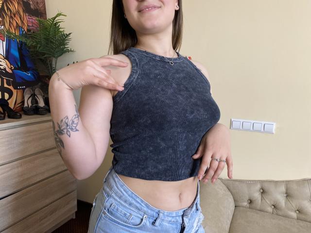 Swoiia, avec une poitrine de taille moyenne, portant un crop top gris foncé, montrant son bras tatoué, et un jean bleu, souriante et touchant son épaule..