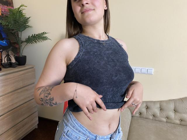 Eine Frau mit heller Haut, braunen Haaren, kleinen Brüsten, einem Tattoo am rechten Arm, die ein dunkles, ärmelloses Oberteil trägt, das sie anhebt und dabei ihren Bauch zeigt, steht in einem Raum, lächelt leicht und trägt blaue Jeans..