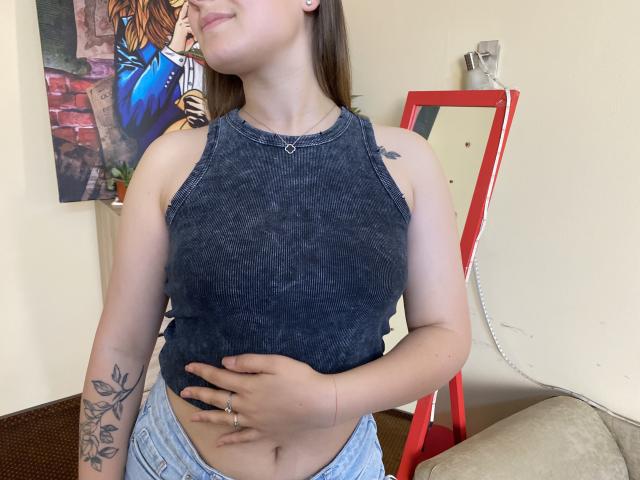 Swoiia, avec de petites seins, portant un haut bleu foncé sans manches, une main sur son ventre, un tatouage sur le bras gauche, debout devant un miroir et une œuvre d'art murale..