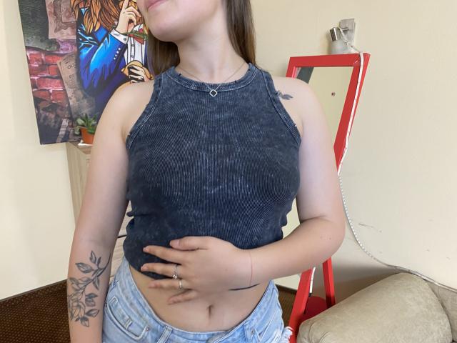 Swoiia vestindo uma cropped sem mangas de cor cinza escura, que mostra seus pequenos seios e a tatuagem em seu braço esquerdo, segurando a barriga, com jeans azuis e cabelo castanho..