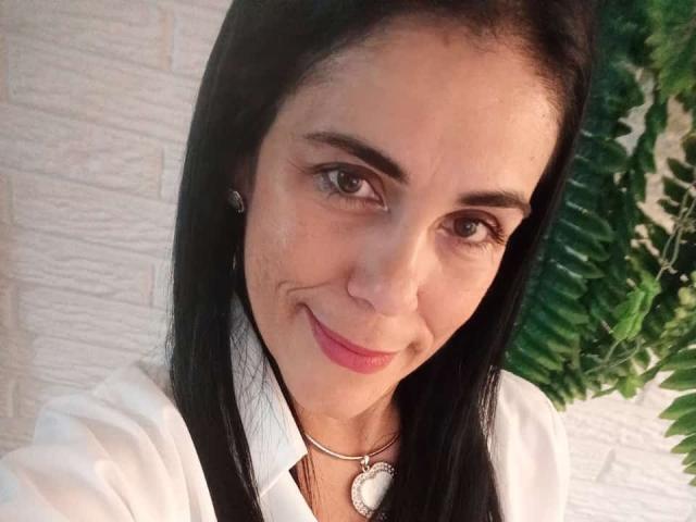GirasolWalton has dark straight hair, brown eyes, and a slight smile with pink lipstick. She's wearing a white shirt and a heart-shaped necklace. Her tits are small.