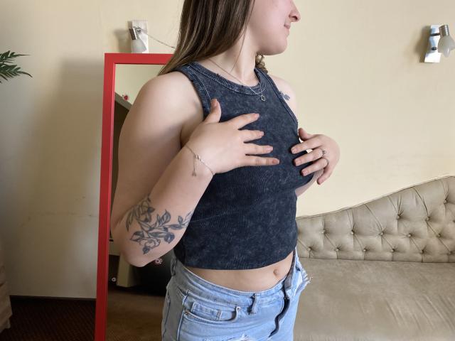 Swoiia, con un seno di media grandezza, indossa una canotta scura, di colore grigio, e jeans azzurri. È in piedi, con le mani sul petto, e mostra un tatuaggio floreale sul braccio destro..