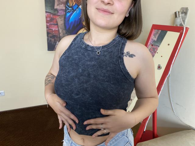 Swoiia, com seios médios, vestindo uma blusa escura cinza, sem mangas e com estampas tie-dye, com as mãos sobre a barriga, shorts jeans, tatuagem no ombro direito, em pé em frente a um espelho..