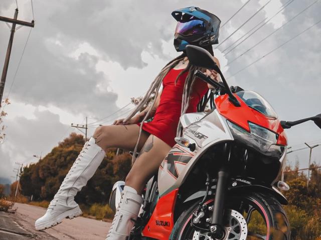 SharlotteX com tranças longas, vestido vermelho, botas brancas, capacete azul, sentada em uma motocicleta vermelha e branca, mostrando uma tatuagem na coxa direita, ao ar livre em um dia nublado..