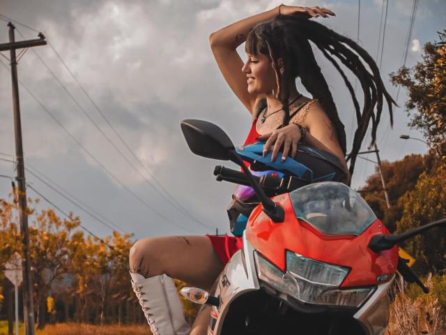 SharlotteX with long black dreads, red tank top, and white lace-up boots, riding a red motorcycle, arm raised, showing off her tits and confident smile.