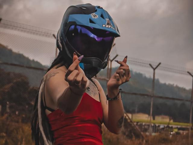 SharlotteX usando um capacete de motocicleta azul com viseira refletiva, apontando com os dedos, tem unhas roxas, vestido vermelho e tatuagens, e está ao ar livre com um céu nublado..