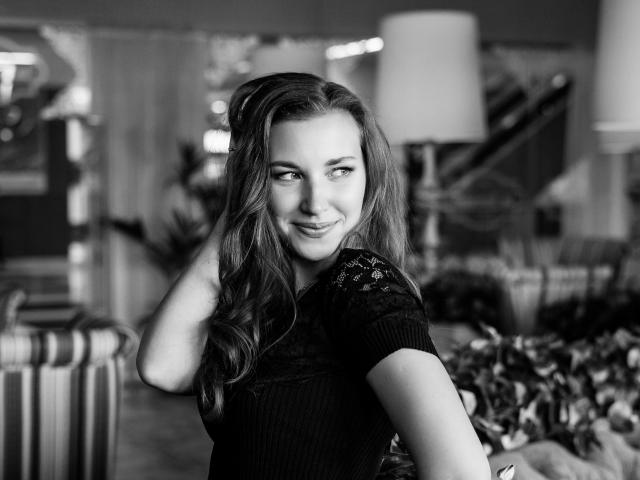 Clarisa, cheveux ondulés, haut en dentelle noire, souriant à la caméra, main dans les cheveux, photo en noir et blanc, montrant discrètement les seins à travers la dentelle, expression douce, décor de chambre, ambiance détendue..