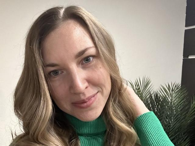 Clarisa, avec ses cheveux blonds ondulés, ses yeux verts et un léger sourire, porte un pull vert à col roulé, sa main derrière sa tête, mettant en valeur son joli nez et ses lèvres roses..