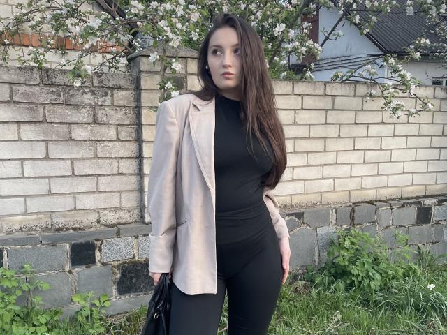 MaryxElla, avec de longs cheveux bruns, portant un blazer rose par-dessus un haut noir moulant et un pantalon noir, tenant un sac noir, debout dans l'herbe, devant un mur de briques..