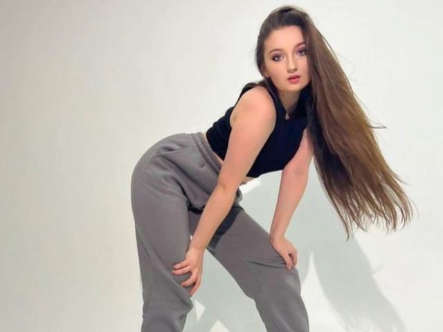 MaryxElla with long brown hair, black sleeveless top, and gray pants, bending forward with hands on knees, looking at camera with slightly parted lips, showing her ass.