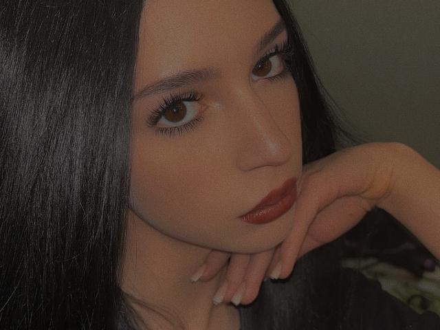AnayaSweety with long black hair, brown eyes, and red lipstick, resting her chin on her hand, showing off her glossy nails, in a dark, moody photo.