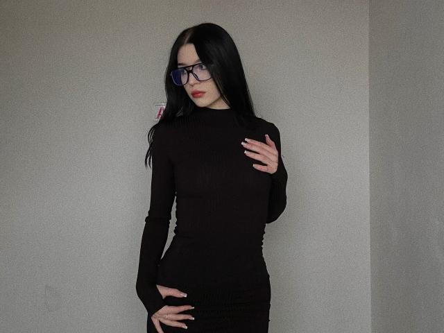 AnayaSweety has long black hair, wearing glasses, a tight black long-sleeve top, and black skirt, touching her chest and hand on her hip, with red lipstick.