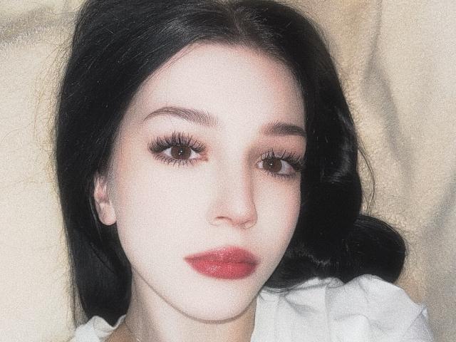 AnayaSweety's face with pale skin, dark black hair, brown eyes, long eyelashes, and red lips. She's wearing a white shirt, looking slightly to the side.