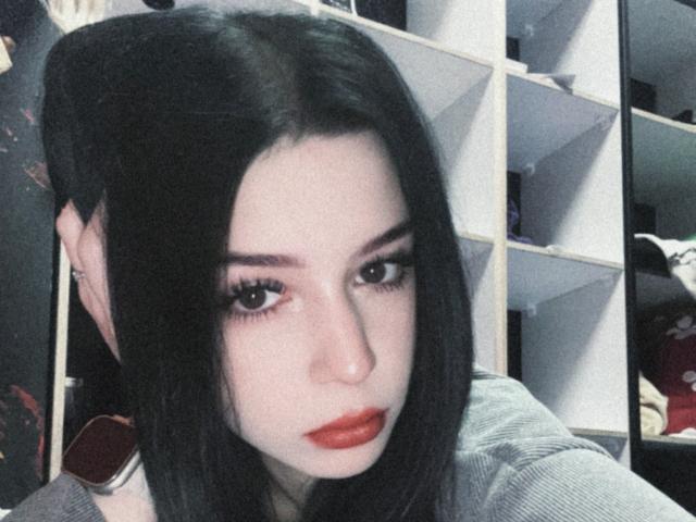 AnayaSweety has pale skin, black straight hair, and red lipstick. She's looking down with half-closed eyes, wearing a gray shirt, in a slightly grainy selfie.